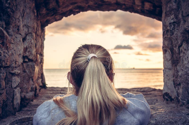 Thoughtful woman devoted into contemplation of beautiful sunset over sea through window of old castle with dramatic sky and perspe