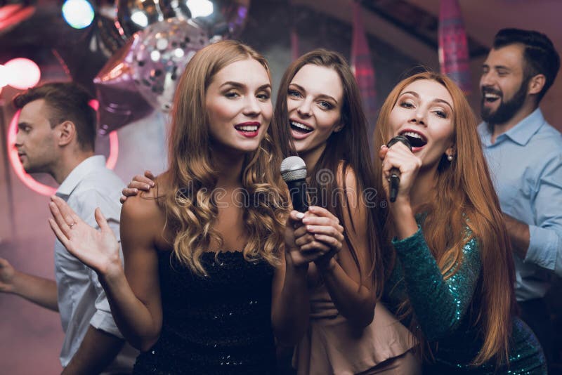 Three beautiful girls sing in a karaoke club. Behind them are men waiting for their turn. Young people have fun in a nightclub.