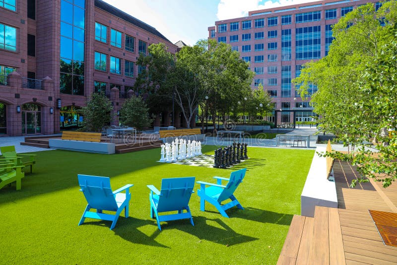 Three blue lawn chairs near a giant chess set on lush green grass in the park surrounded by lush green trees and buildings