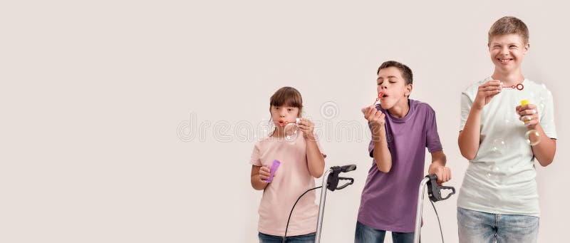 Three cheerful disabled children with Down syndrome and cerebral palsy smiling while blowing soap bubbles, standing