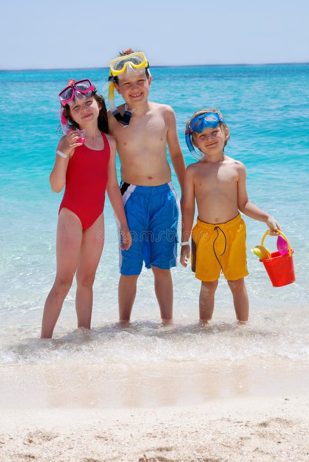 Three siblings stand together with ocean water in background. One girl and two boys are wearing swimsuits and snorkeling gear. Youngest male child is carrying pail. Three siblings stand together with ocean water in background. One girl and two boys are wearing swimsuits and snorkeling gear. Youngest male child is carrying pail.