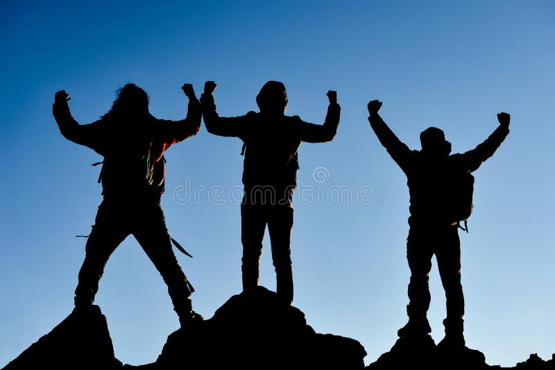 Team of three successful climbers celebrating upon reaching the mountain summit seen in silhouette against a pale blue sky. Team of three successful climbers celebrating upon reaching the mountain summit seen in silhouette against a pale blue sky.