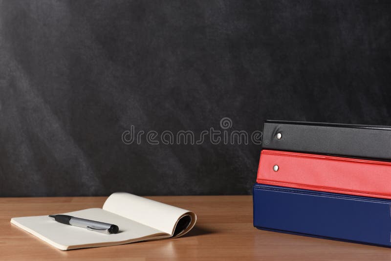 Three different binders with note pad and pen laying on a teachers desk in front of a chalk board