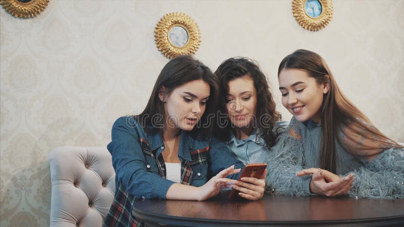 Three friends, sitting in a coffee shop using a smart phone and having a funny conversation. Good girls with long