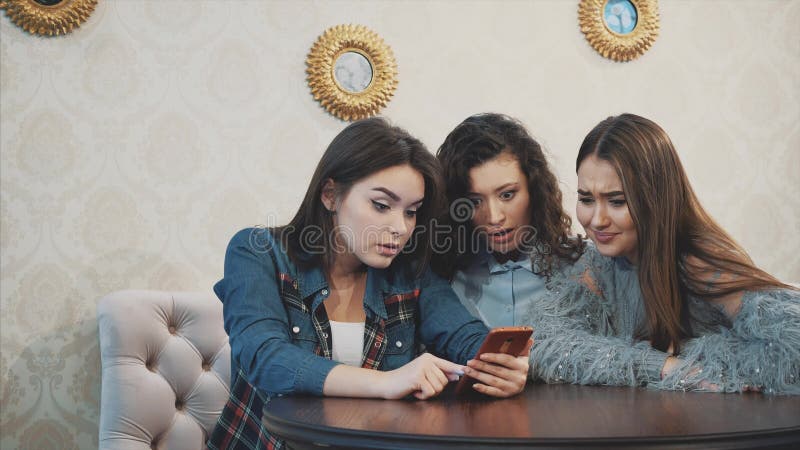 Three friends, sitting in a coffee shop using a smart phone and having a funny conversation. Good girls with long