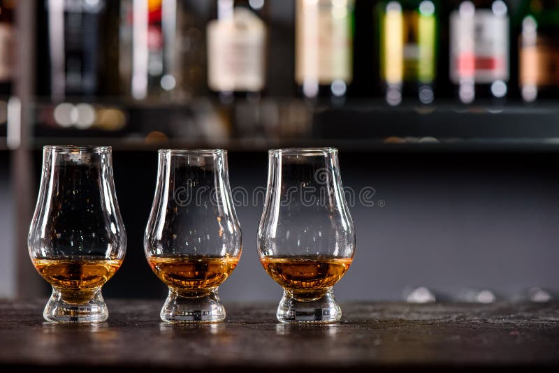 Three Glencairn Glass with whiskey on a bar wooden counter close up on the background of blurry bottles