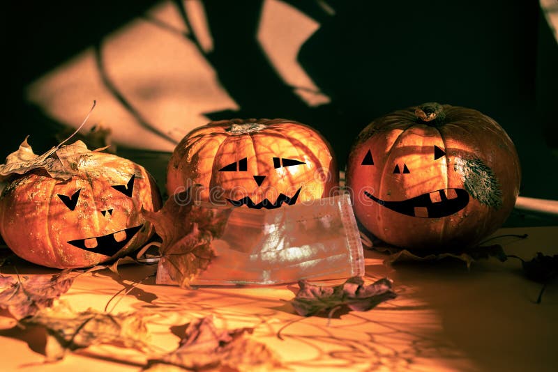 Three halloween pumpkins painted as Jack`s lantern, one in protective face mask, fall leaves, magic shadows. New normal stock photo