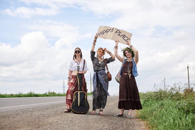 Three hippie women, wearing boho style clothes, standing on road, thumbing a ride, hitchhiking with sign Happy place on cardboard. Friends, traveling together in summer. Freedom and happiness concept