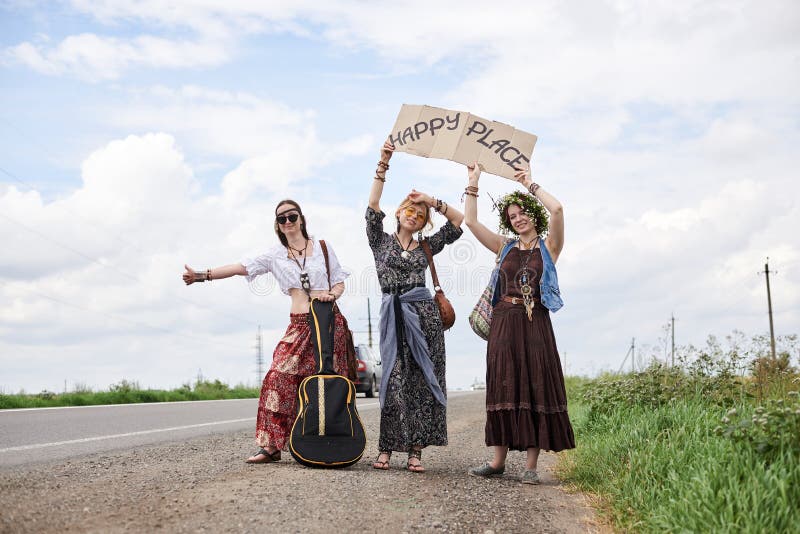 Three hippie women, wearing boho style clothes, standing on road, thumbing a ride, hitchhiking with sign Happy place on cardboard. Friends, traveling together in summer. Freedom and happiness concept