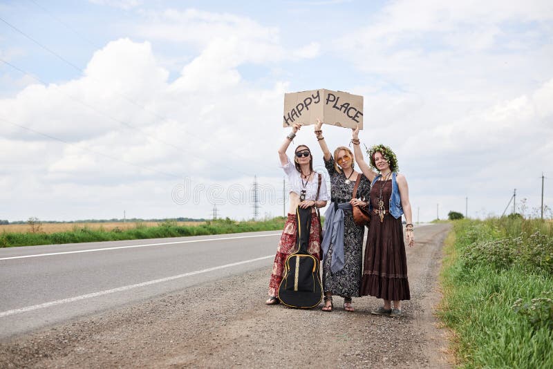 Three hippie women, wearing boho style clothes, standing on road, thumbing a ride, hitchhiking with sign Happy place on cardboard. Friends, traveling together in summer. Freedom and happiness concept