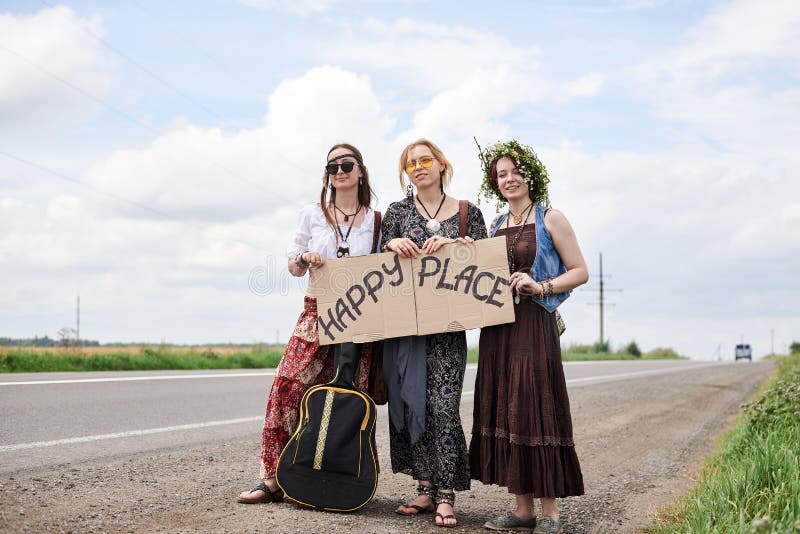 Three hippie women, wearing boho style clothes, standing on road, thumbing a ride, hitchhiking with sign Happy place on cardboard. Friends, traveling together in summer. Freedom and happiness concept