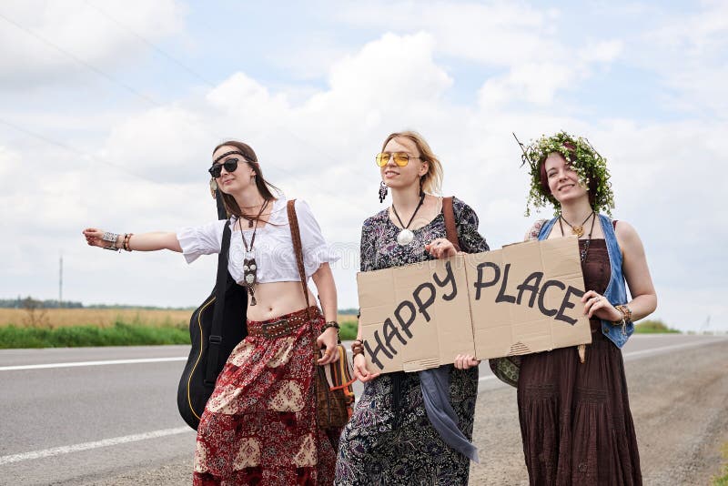 Three hippie women, wearing boho style clothes, standing on road, thumbing a ride, hitchhiking with sign Happy place on cardboard. Friends, traveling together in summer. Freedom and happiness concept