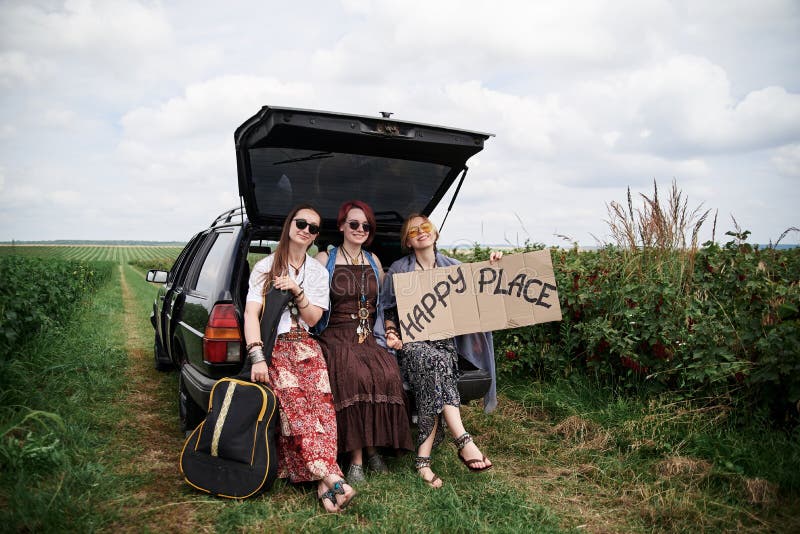 Three hippie women, wearing colorful boho style clothes, sitting on car trunk, holding Happy place sign, smiling, relaxing. Friends, traveling together in rural countryside. Eco tourism concept
