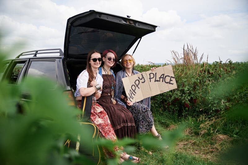 Three hippie women, wearing colorful boho style clothes, sitting on car trunk, holding Happy place sign, smiling, relaxing. Friends, traveling together in rural countryside. Eco tourism concept