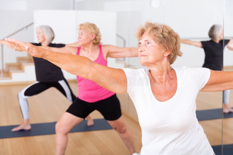 Three mature women doing yoga in a group class perform the exercise by taking the warrior pose II. Three mature women doing yoga in a group class perform the exercise by taking the warrior pose II