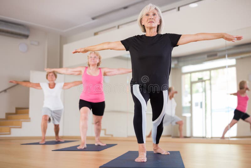 Three mature women doing yoga in a group class perform the exercise by taking the warrior pose II. Three mature women doing yoga in a group class perform the exercise by taking the warrior pose II