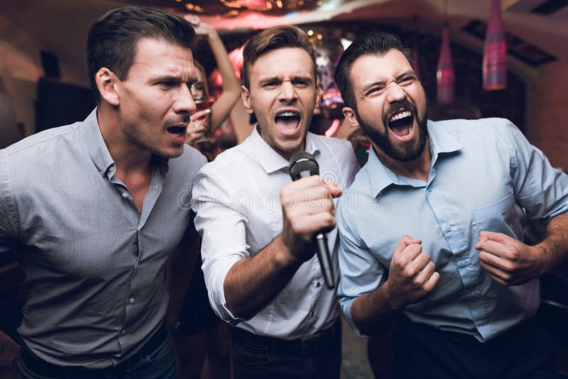 Three men sing at a karaoke club. Young people have fun in a nightclub. They are very cheerful and they smile.
