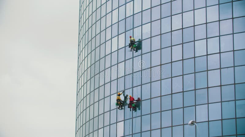 Three men workers hanging on ropes and cleaning the exterior glass windows of a business skyscraper after rain - autumn