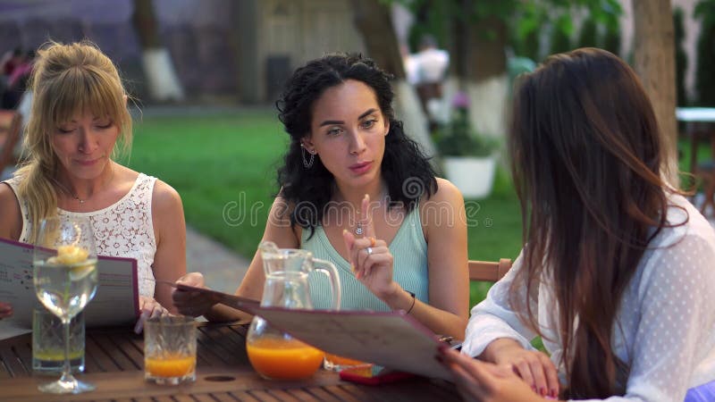 Three pretty girlfriends talking and reading menu in cafe outdoors in the park stock video