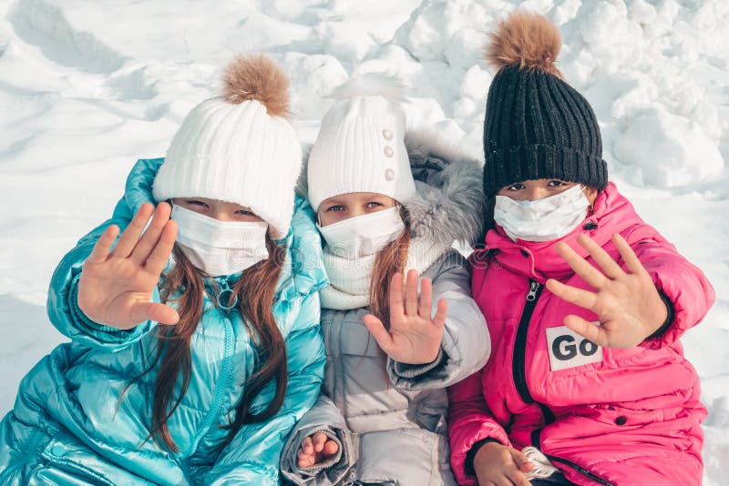 Three teenage girls in protective masks are sitting on the snow in the park. Girls show a gesture of denial. Stop the epidemic of viruses and flu. Stop coronovirus.