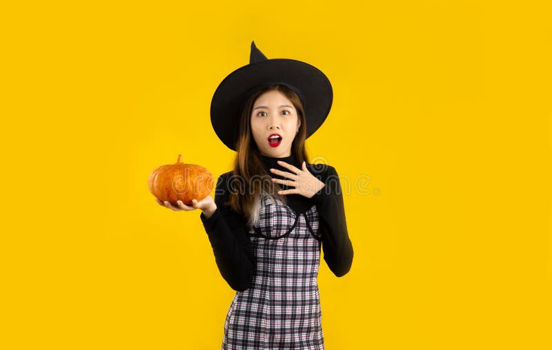 Halloween theme, young asian woman in black dress wearing witch hat holding orange pumpkin posing surprised and excited on yellow backgroud. Halloween theme, young asian woman in black dress wearing witch hat holding orange pumpkin posing surprised and excited on yellow backgroud.