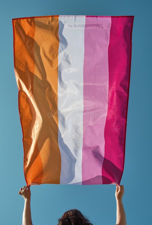 Closeup of a caucasian woman outdoors holding a lesbian pride flag above her head, moved by the air, against the sky. Closeup of a caucasian woman outdoors holding a lesbian pride flag above her head, moved by the air, against the sky