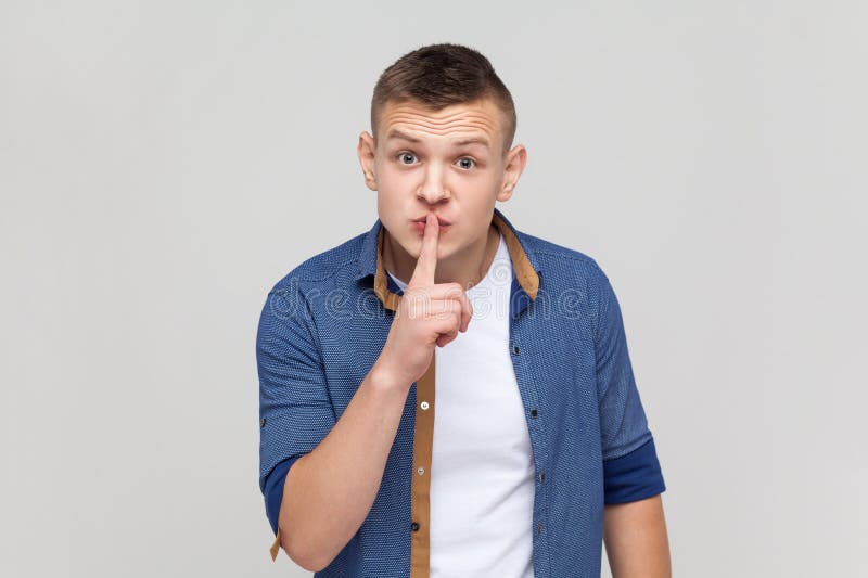 Please, be quiet. Teenager boy in blue shirt shushing with silence gesture, holding finger on lips and smiling to camera, asking to keep secret. Indoor studio shot isolated on gray background. Please, be quiet. Teenager boy in blue shirt shushing with silence gesture, holding finger on lips and smiling to camera, asking to keep secret. Indoor studio shot isolated on gray background.