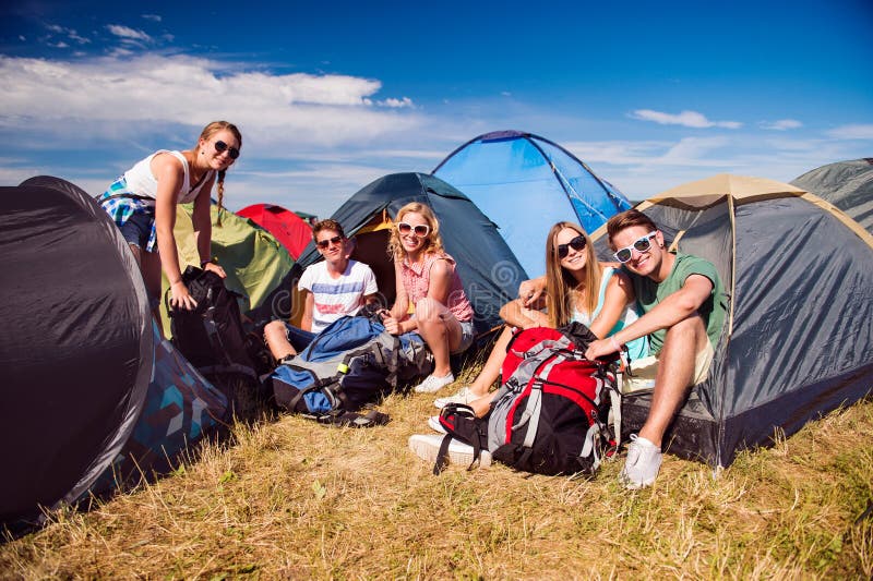 Group of teenage boys and girls at summer music festival, sitting on the ground in front of tents, packing. Group of teenage boys and girls at summer music festival, sitting on the ground in front of tents, packing