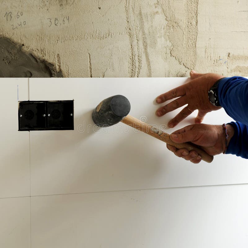 Tiling the kitchen with white porcelain stoneware tiles. Square format image. A person wearing a blue long-sleeve shirt uses a rubber mallet to adjust a white tile. The tile is being positioned near an electrical outlet box set in a wall with a rough, unfinished plaster surface. The scene suggests tile installation, typically part of a renovation process. Handwritten measurements are visible on the wall above the tile. Bricklayer tiler stock images, royalty-free photos and pictures