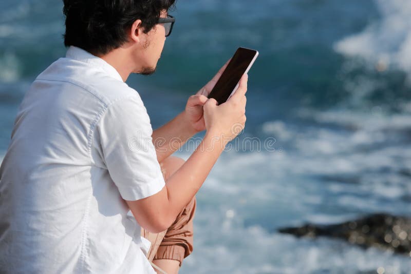 Tillbaka sikt av den unga asiatiska mannen som använder den mobila smarta telefonen på havskusten Internet av sakerbegreppet arkivbild Tillbaka sikt av den unga asiatiska mannen som använder den mobila smarta telefonen på havskusten Internet av sakerbegreppet arkivbild