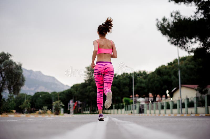Tillbaka sikt av spring för ung kvinna längs huvudvägen i morgonen. Tillbaka sikt av iklädd rosa sportswearspring för ung kvinna längs huvudvägen i morgonen