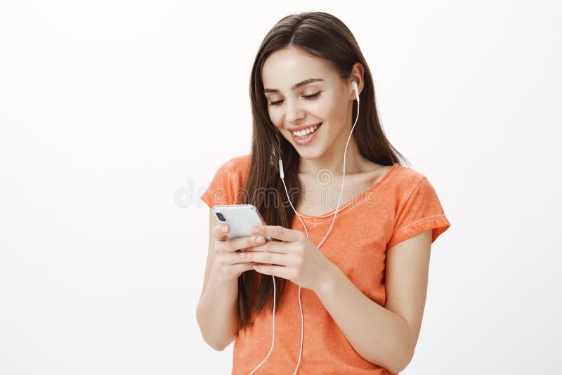 Tillfredsställd attraktiv blyg flicka i earbuds, lyssnande musik som väljer det favorit- spåret i smartphoneplaylisten som ler fotografering för bildbyråer Tillfredsställd attraktiv blyg flicka i earbuds, lyssnande musik som väljer det favorit- spåret i smartphoneplaylisten som ler fotografering för bildbyråer
