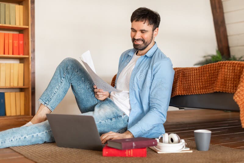 A cheerful man in casual attire sits on the floor with his laptop open and paperwork in hand, sitting area features a coffee cup, headphones, and books, suggesting a relaxing work-from-home setup. A cheerful man in casual attire sits on the floor with his laptop open and paperwork in hand, sitting area features a coffee cup, headphones, and books, suggesting a relaxing work-from-home setup