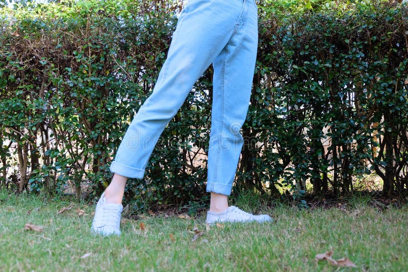 Tillfälliga vita gymnastikskor för Hipster Kvinnliga stående bärande vitskor och jeans flåsar på naturbakgrund för grönt gräs arkivbilder Tillfälliga vita gymnastikskor för Hipster Kvinnliga stående bärande vitskor och jeans flåsar på naturbakgrund för grönt gräs arkivbilder
