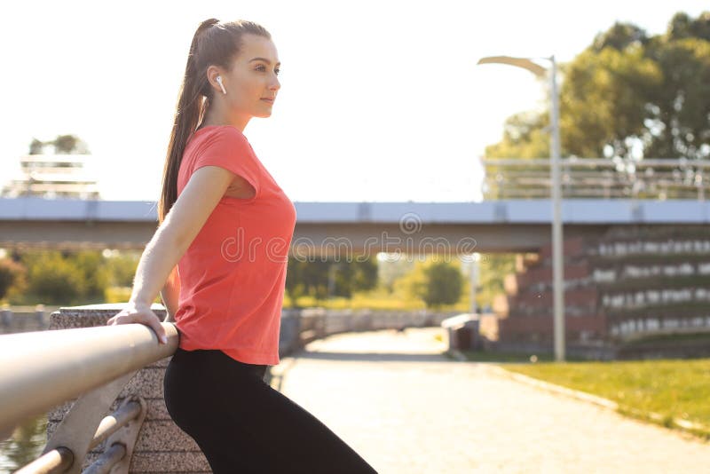 Tired attractive young woman in sports clothing relaxing while standing outdoors stock photo Tired attractive young woman in sports clothing relaxing while standing outdoors stock photo