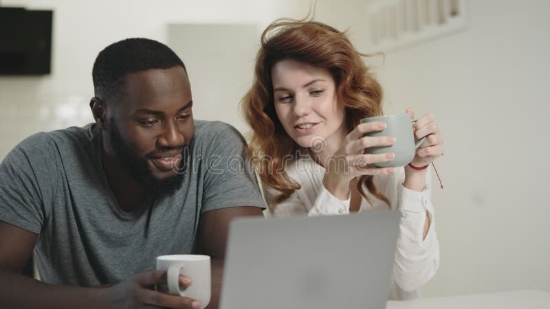 Tired black man looking laptop screen at home. Woman bringing tea to guy.