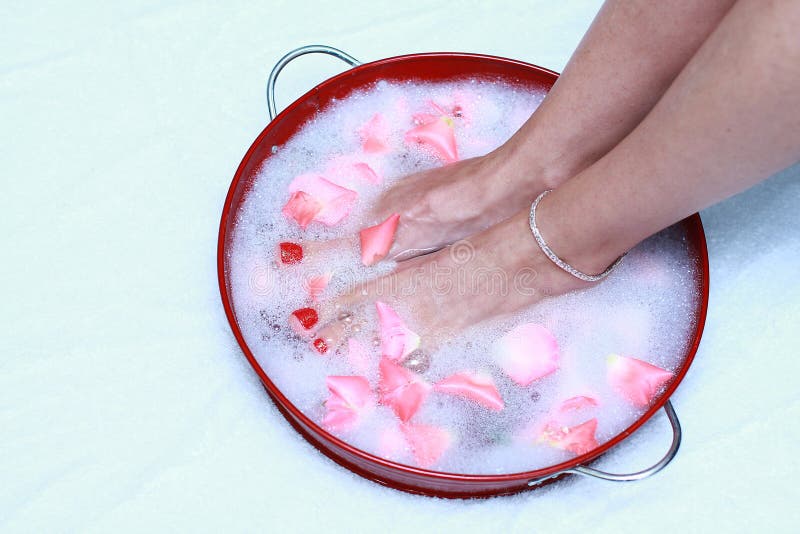 A Caucasian woman's feet soak in a red pan of warm, bubbly water with rose petals floating. She wears red toenail polish and a silver ankle bracelet. A Caucasian woman's feet soak in a red pan of warm, bubbly water with rose petals floating. She wears red toenail polish and a silver ankle bracelet.