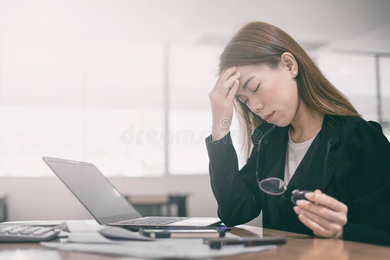Tired and headache asian business woman closed eyes and holding glasses sitting at desk in front of laptop at office background stock images Tired and headache asian business woman closed eyes and holding glasses sitting at desk in front of laptop at office background stock images