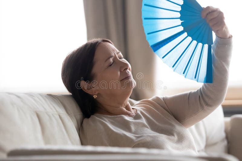 Tired middle aged woman with closed eyes waving blue paper fan, suffering from heat, sitting on couch at home, sweaty mature female feeling unwell, cooling in hot summer weather, high temperature. Tired middle aged woman with closed eyes waving blue paper fan, suffering from heat, sitting on couch at home, sweaty mature female feeling unwell, cooling in hot summer weather, high temperature