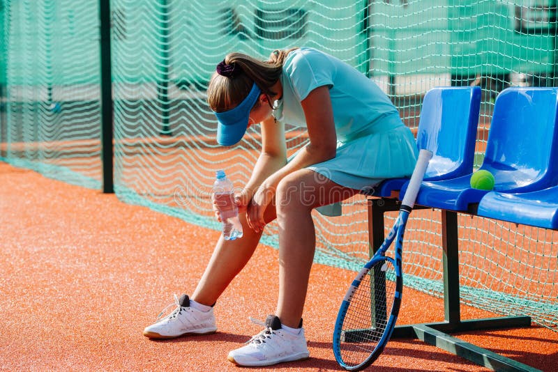 Tired teenage girl sitting on a chair bench next to tennis court to take a short break