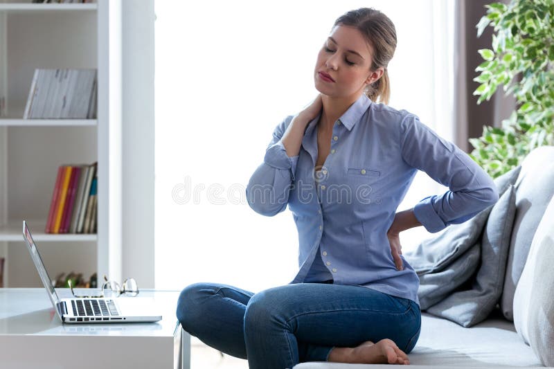 Tired young woman with shoulder and back pain sitting on the couch at home. stock photo