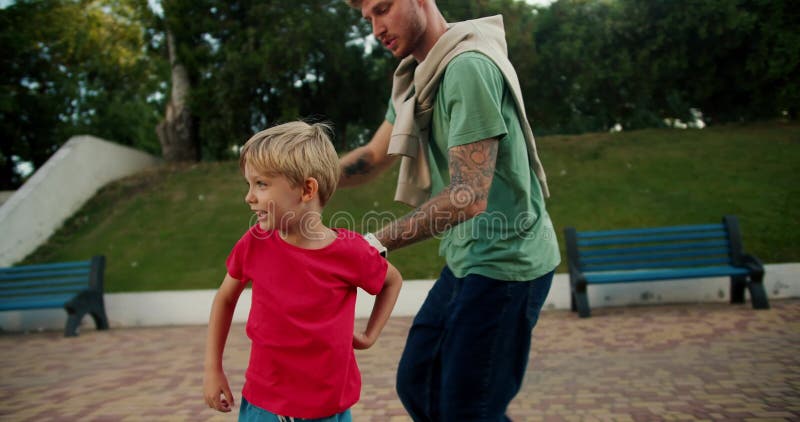 Tiro em tela de um pai de jeans azuis ajudando seu filho em uma camisa vermelha andando de skate no parque. pouco