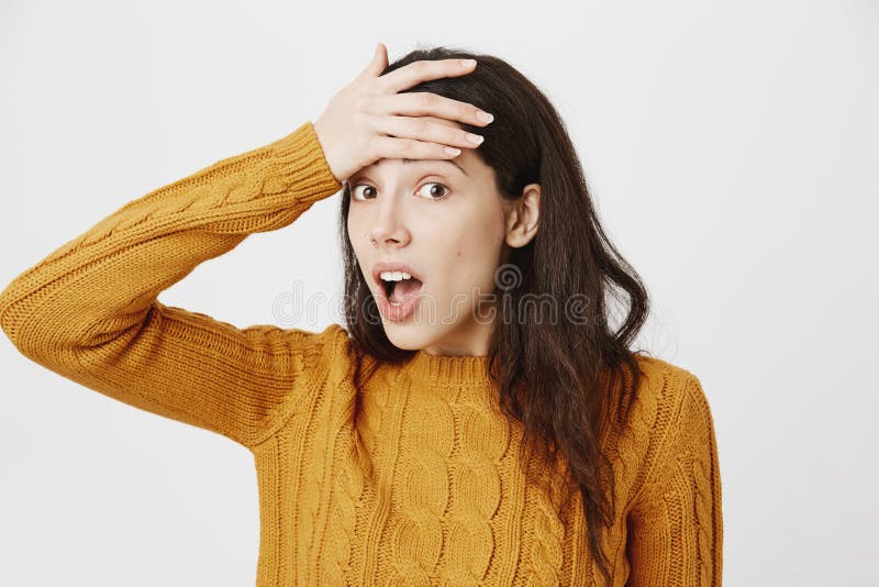 Tiro interior de la muchacha europea desconcertada y olvidadiza, llevando a cabo la mano en la frente y la situación con la boca imágenes de archivo libres de regalías Tiro interior de la muchacha europea desconcertada y olvidadiza, llevando a cabo la mano en la frente y la situación con la boca imágenes de archivo libres de regalías