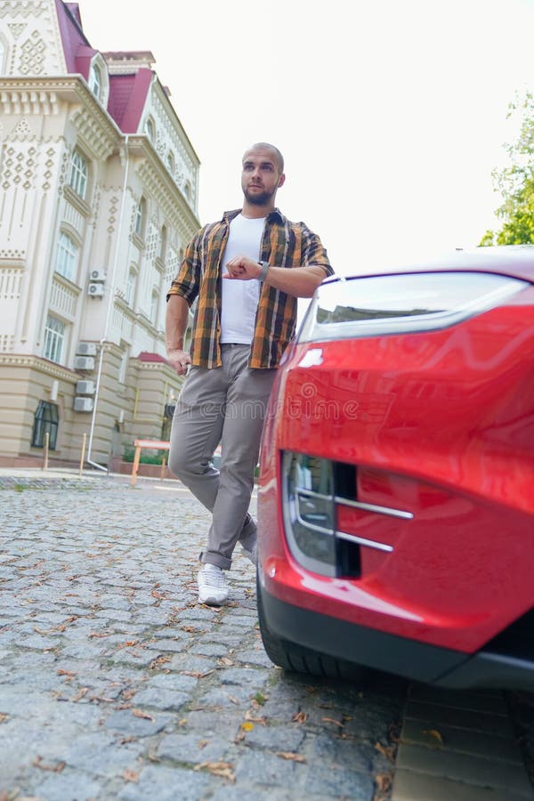 Tiro vertical del individuo barbudo intrépido apuesto joven que se coloca al aire libre cerca de su coche rojo que comprueba el t imagen de archivo Tiro vertical del individuo barbudo intrépido apuesto joven que se coloca al aire libre cerca de su coche rojo que comprueba el t imagen de archivo