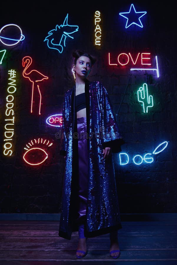 Cyberpunk style portrait of girl in futuristic purple bathrobe with glitter. She poses against wall of neon figures. Set is lit with magenta light. Clothes is oversized. Picture has dark noir tones. Cyberpunk style portrait of girl in futuristic purple bathrobe with glitter. She poses against wall of neon figures. Set is lit with magenta light. Clothes is oversized. Picture has dark noir tones.