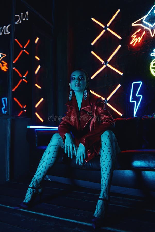 Cyberpunk style portrait of girl in futuristic red bikers jacket and tights in net. She poses on leather sofa against neon figures. Set is lit with blue light from TV. Picture has dark noir tones. Cyberpunk style portrait of girl in futuristic red bikers jacket and tights in net. She poses on leather sofa against neon figures. Set is lit with blue light from TV. Picture has dark noir tones.