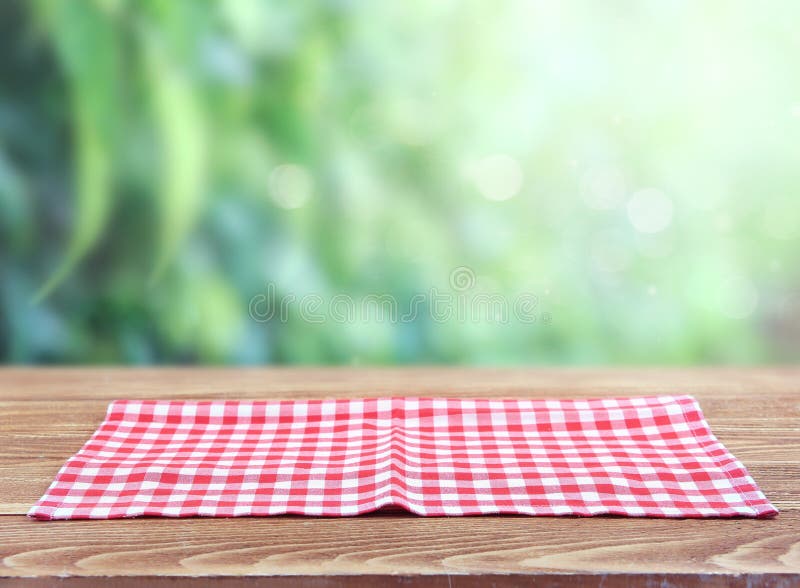 Checkered red picnic folded towel cloth on wooden table blurred green natural background.Food advertisement  backdrop. Checkered red picnic folded towel cloth on wooden table blurred green natural background.Food advertisement  backdrop