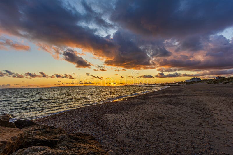 Titel: En bild av en stonstrand vid solnedgången med krasande vågor och eko under en stor gul grumlig himmel arkivfoton