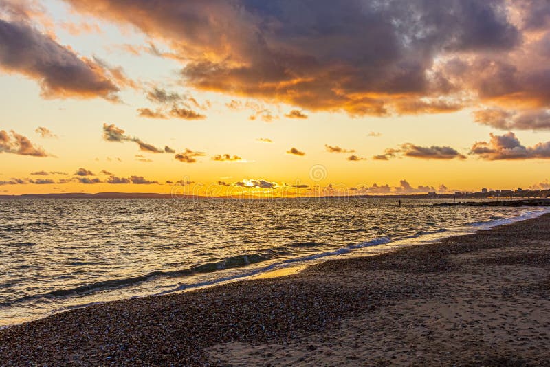Titel: En bild av en stonstrand vid solnedgången med krasande vågor och eko under en stor gul grumlig himmel arkivfoton