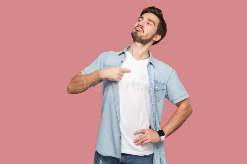 This is me. Portrait of proud haughty handsome bearded young man in blue casual style shirt standing, looking away and pointing himself. indoor studio shot,  on pink background. This is me. Portrait of proud haughty handsome bearded young man in blue casual style shirt standing, looking away and pointing himself. indoor studio shot,  on pink background